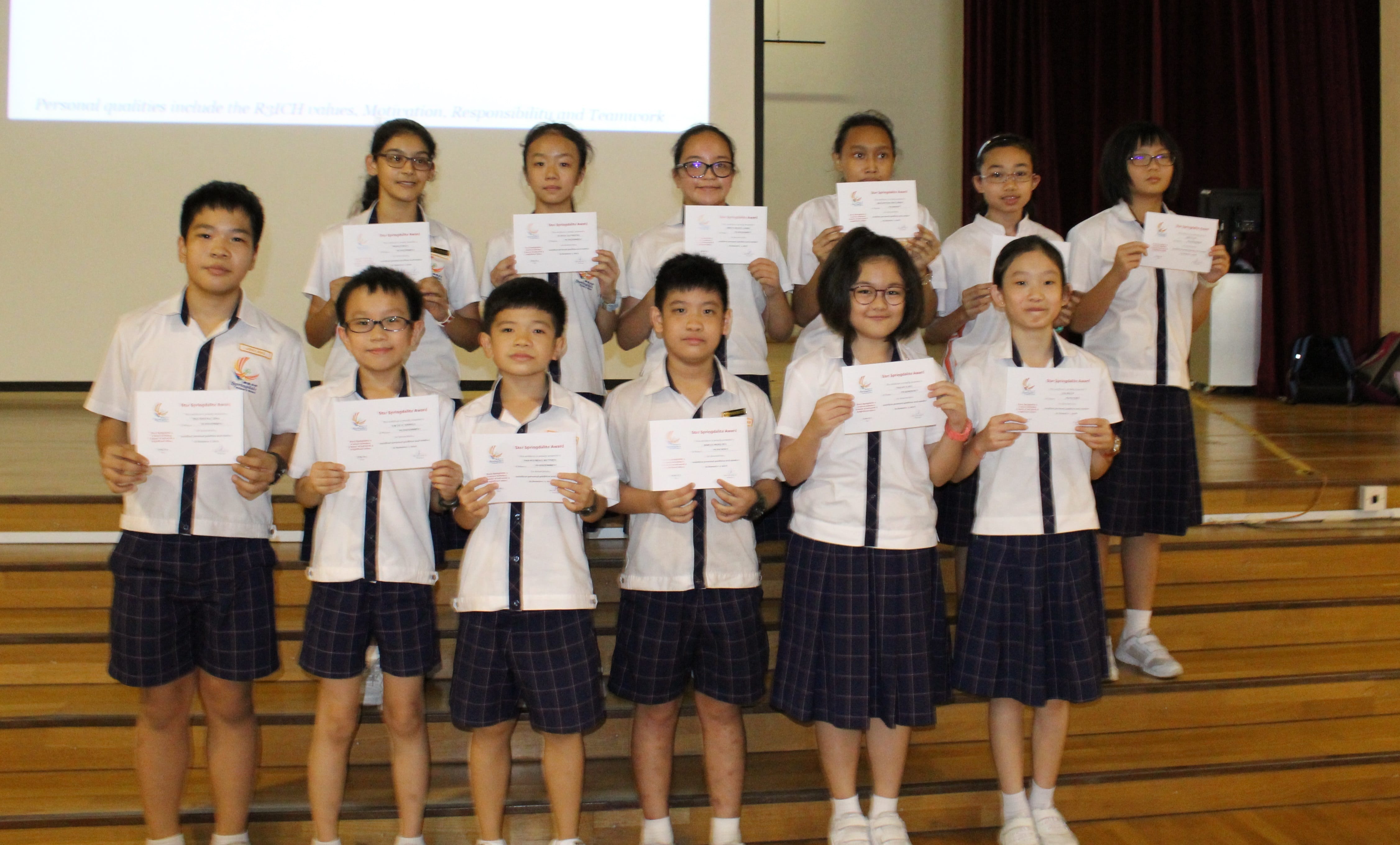 Twelve school children in white uniforms pose on stage steps, each holding a certificate award.